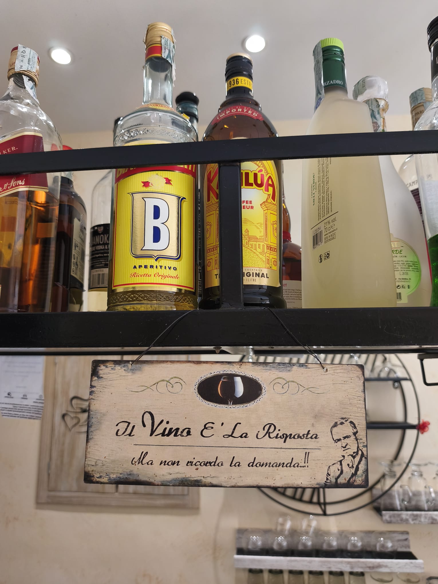 Mensole di un bar con diverse bottiglie di liquore e un pittoresco cartello in legno