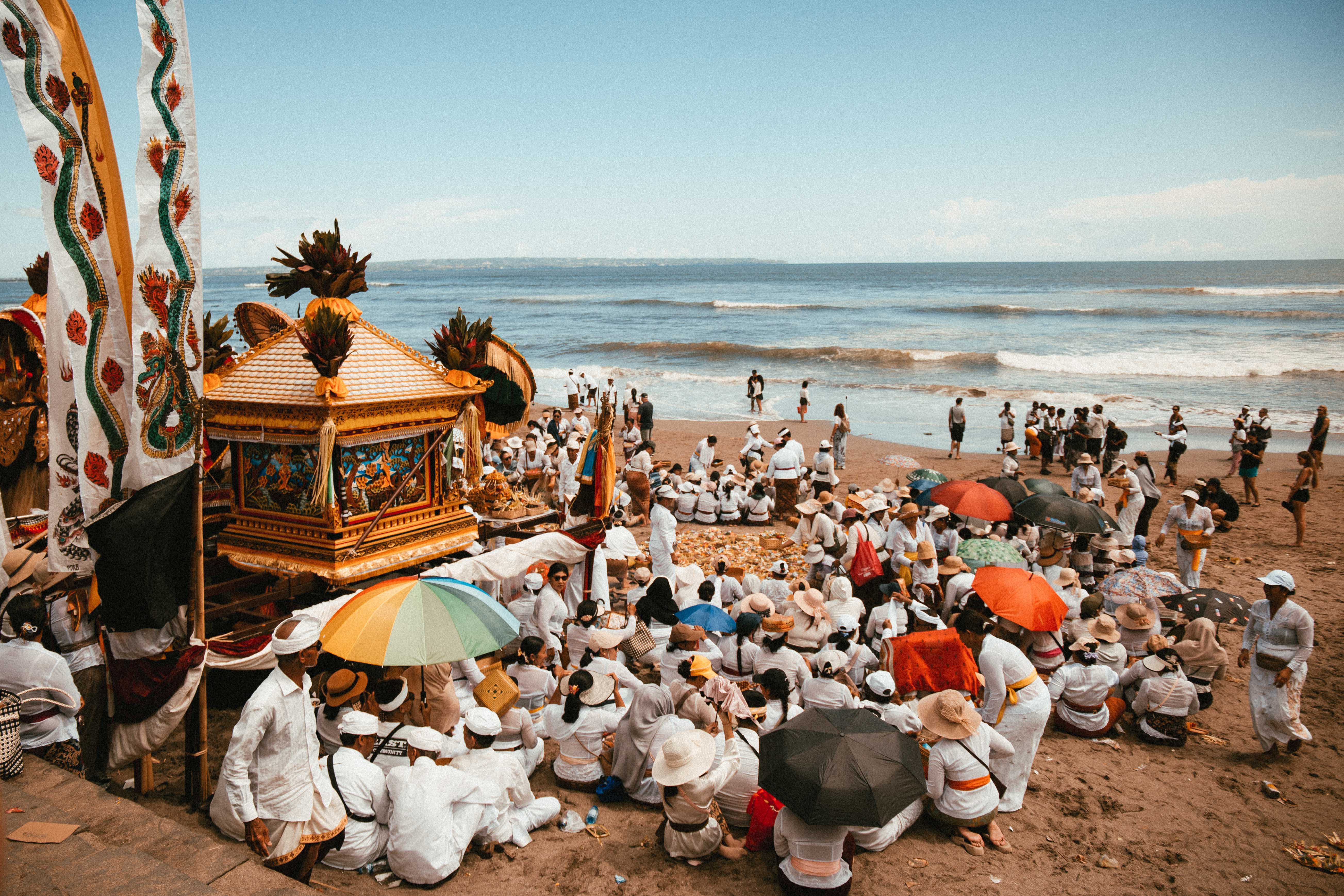 Una grande cerimonia balinese sulla spiaggia, fedeli seduti sulla sabbia con offerte e ombrelli tradizionali bianchi e colorati